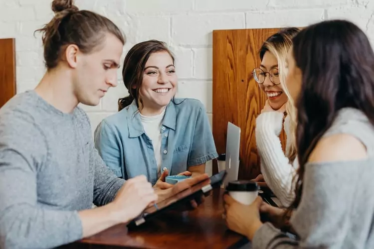 four people at a table with coffee talking and smiling while looking at a laptop and a tablet