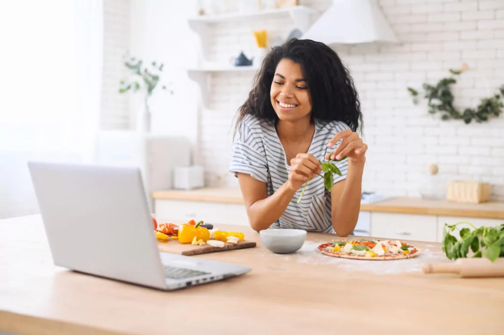 a woman in her kitchen preparing a pizza and looking at a laptop