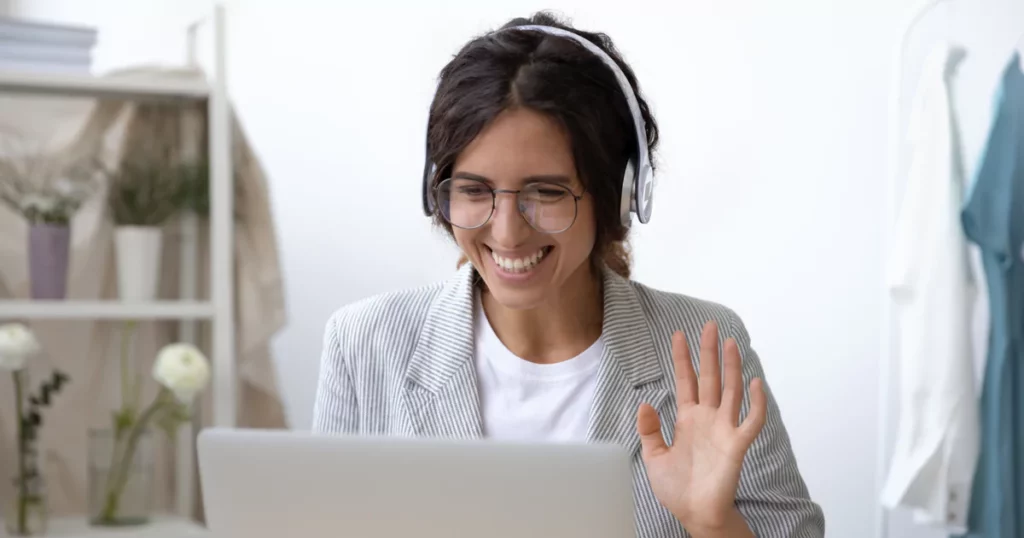 woman with headphones and blazer smiling and waving at a laptop